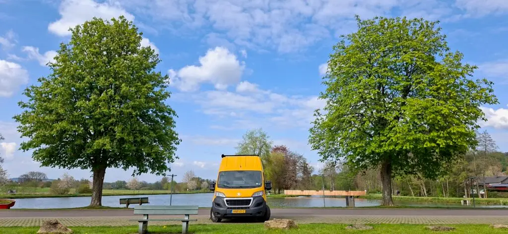 “Gele Happy Vanlife offgrid camper geparkeerd tussen twee grote bomen aan een rustig meer, onder een blauwe lucht met wolken.”