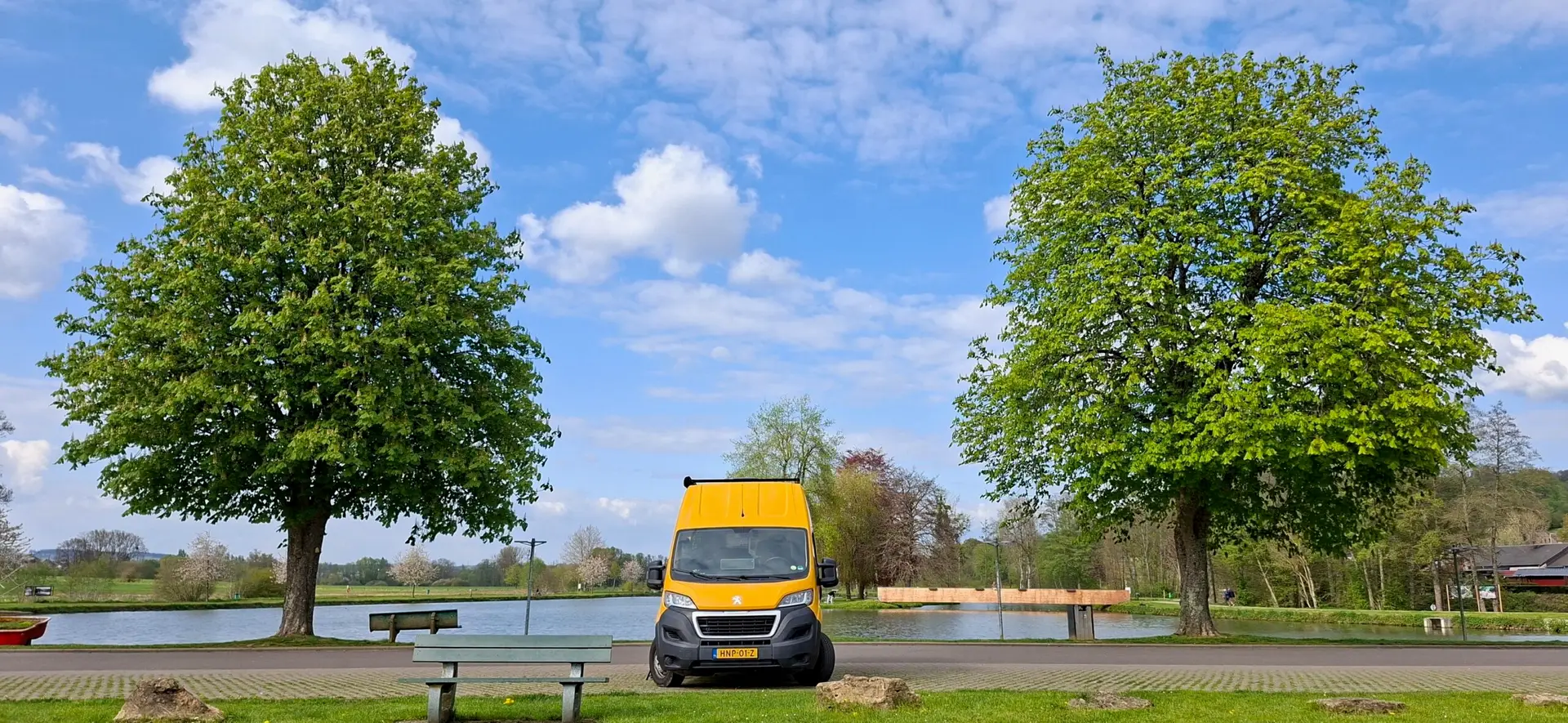 “Gele Happy Vanlife offgrid camper geparkeerd tussen twee grote bomen aan een rustig meer, onder een blauwe lucht met wolken.”