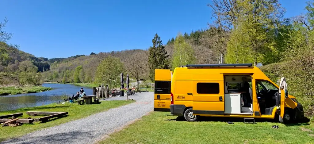 “Happy Vanlife offgrid camperbus geparkeerd aan een rivier op een groene natuurcamping, met volledig uitgeruste camper voor avontuurlijk reizen in de natuur.”
