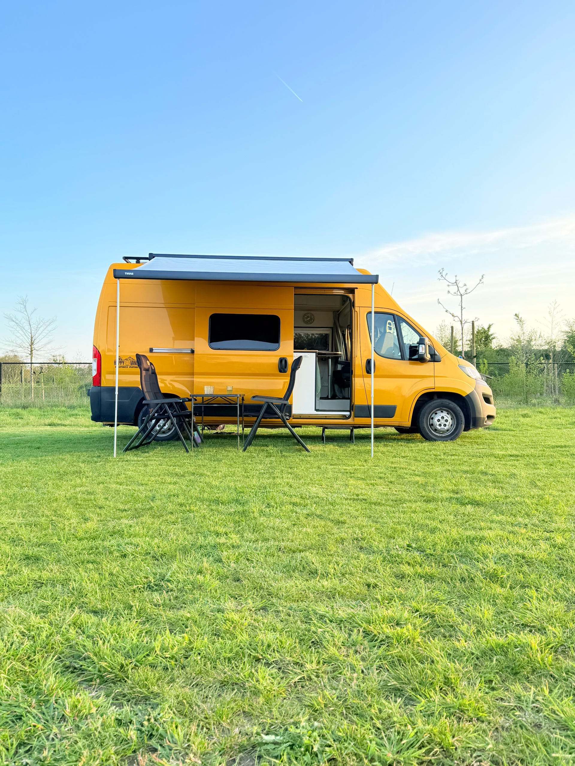 Gele off-grid campervan van Happy Vanlife met luifel, tafel en stoelen op een groen grasveld onder een heldere blauwe lucht.