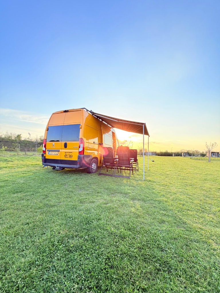 Gele off-grid campervan van Happy Vanlife op een uitgestrekt grasveld tijdens zonsondergang met luifel en campingstoelen buiten.