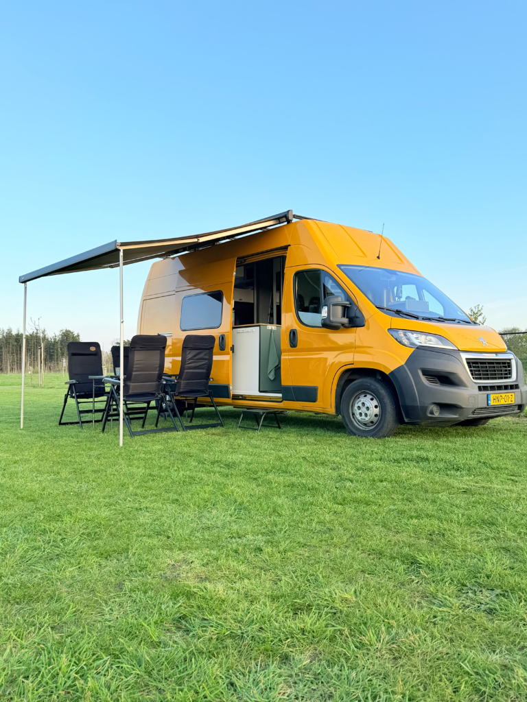 Gele off-grid campervan van Happy Vanlife met luifel en campingstoelen op een groen veld onder een heldere blauwe lucht.