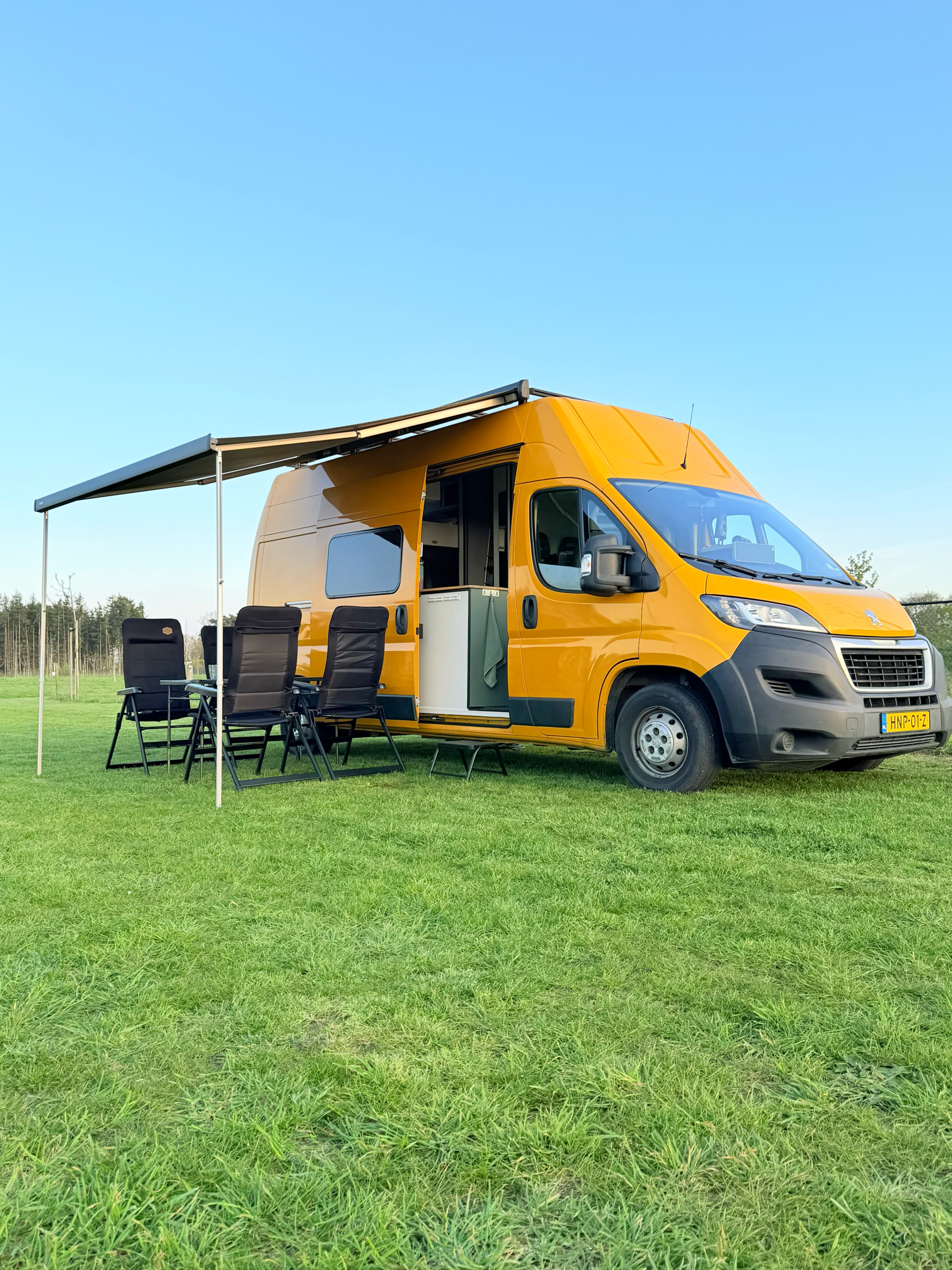 Gele off-grid campervan van Happy Vanlife met luifel en campingstoelen op een groen veld onder een heldere blauwe lucht.